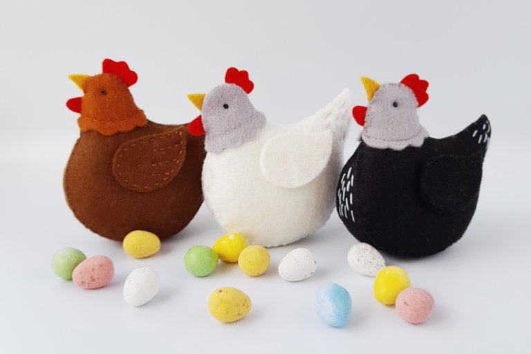 Three handsewn felt hens , one brown, one white and one black sitting with easter eggs around them, on a white background.